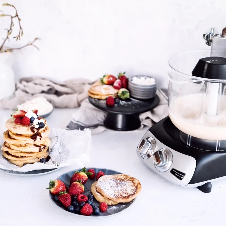 Ankarsrum Assistent Original stand mixer beside stacks of fluffy pancakes topped with fresh strawberries, raspberries, and blueberries.