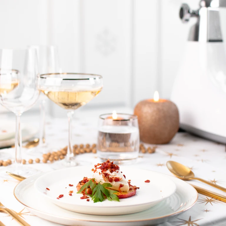 Elegant holiday appetizer plate with whipped cream topping, herbs, and crispy garnish beside white wine glasses and candles.