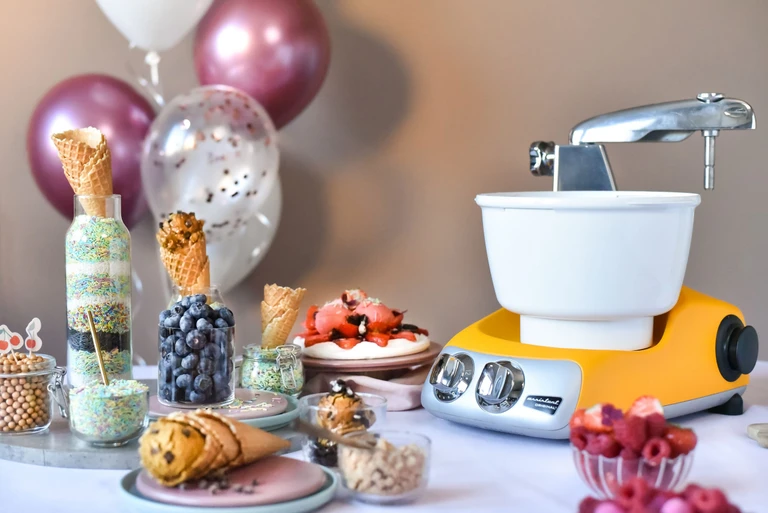 Ankarsrum Original Stand Mixer in yellow with ice cream bowl attachment surrounded by ice cream toppings, cones, blueberries, raspberries, and sprinkles on a festive table.
