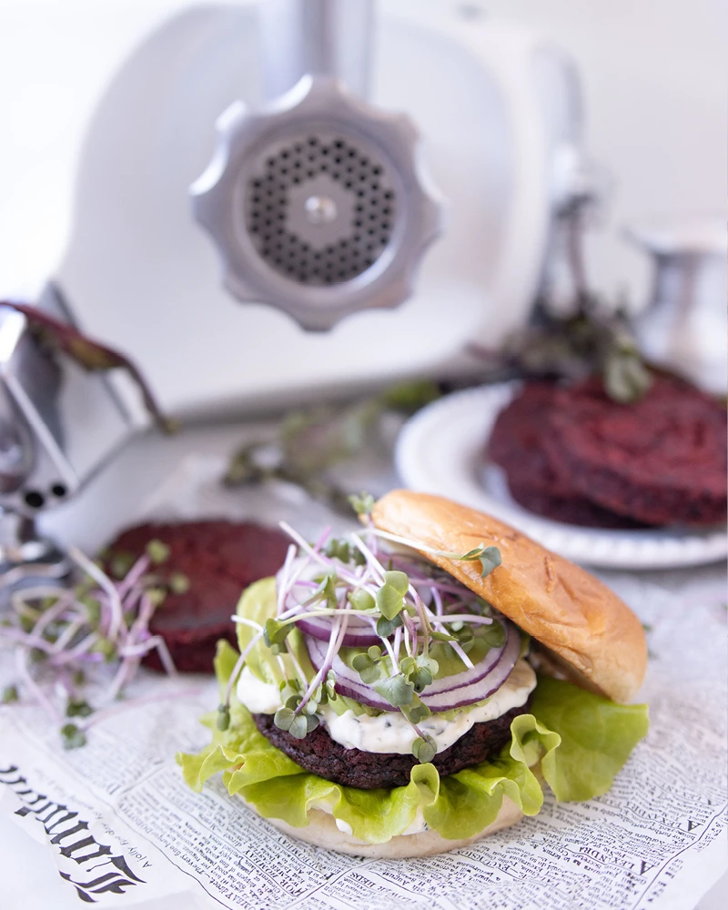 Ankarsrum stand mixer with meat grinder attachment beside a homemade beet burger with lettuce, red onion, and microgreens.