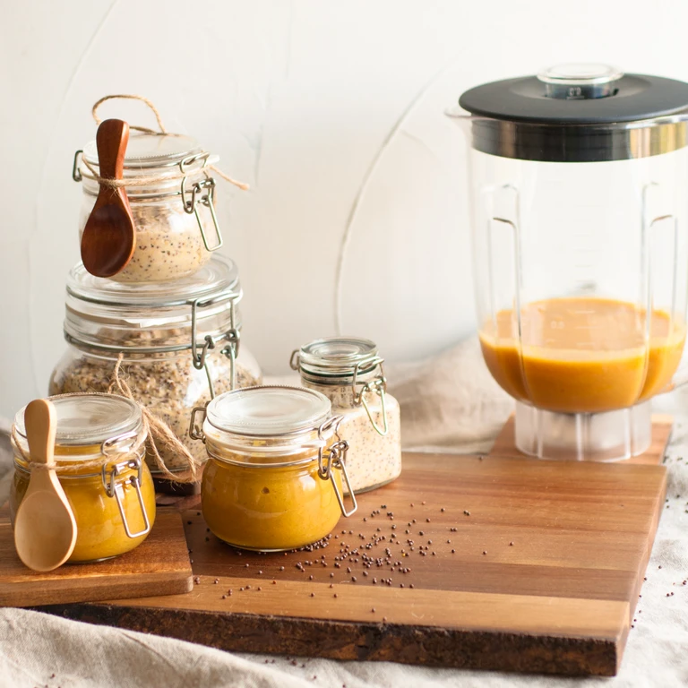 Ankarsrum blender filled with golden sauce beside glass jars of mustard, grains, and seeds on a wooden cutting board.