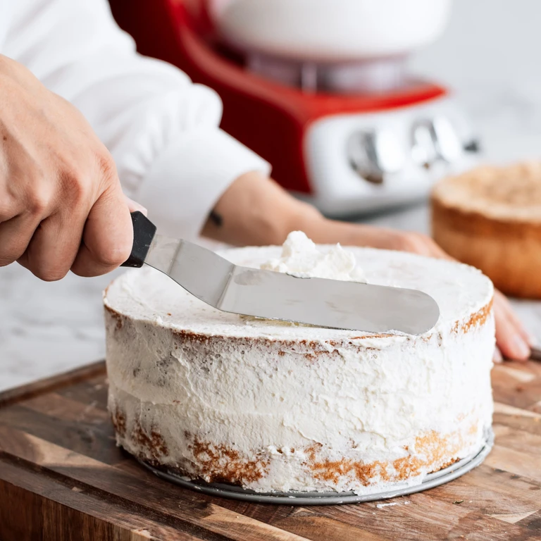 Baker applying white frosting to a layered cake using an offset spatula, with a red Ankarsrum stand mixer in the background.
