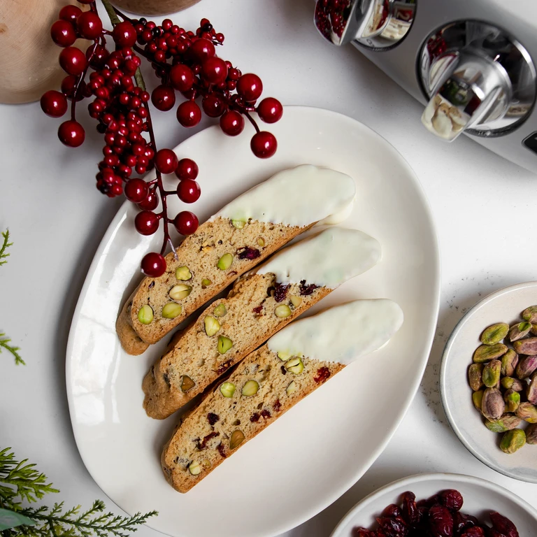 Three white chocolate-dipped pistachio cranberry biscotti on a white oval plate with festive red berry decoration.