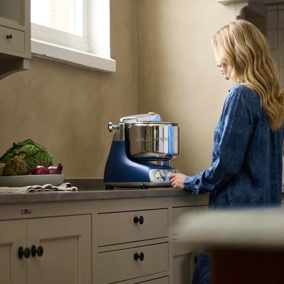 Ankarsrum Assistent Original in Royal Blue in a kitchen, with a woman standing in front of the machine.