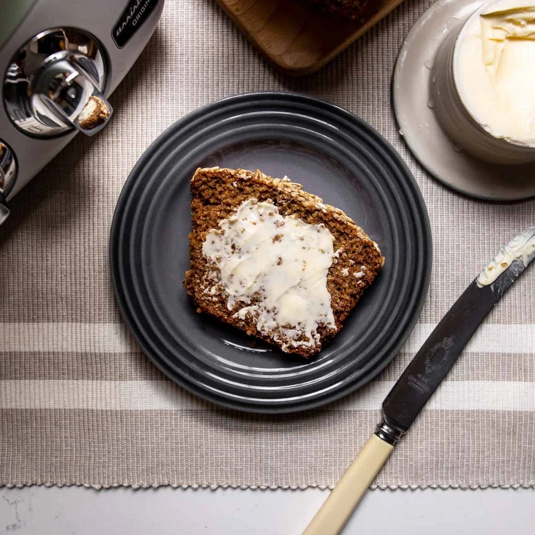 Slice of homemade oat bread with butter on a dark plate, made with an Ankarsrum Original stand mixer nearby.