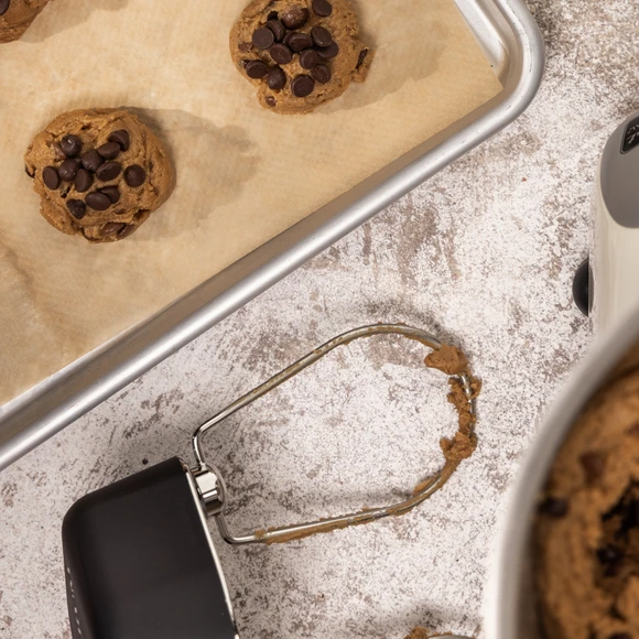 Cookie dough balls with chocolate chips on parchment-lined baking sheet, cookie scoop, and Ankarsrum mixer bowl nearby.