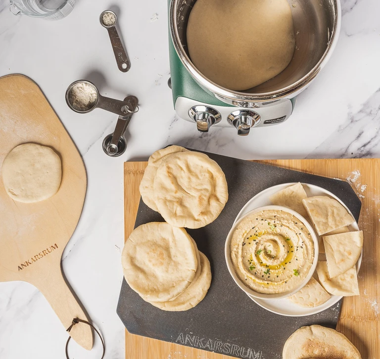 Ankarsrum stand mixer with pita bread dough, baked pitas, hummus dip, measuring spoons, and branded wooden peel board.