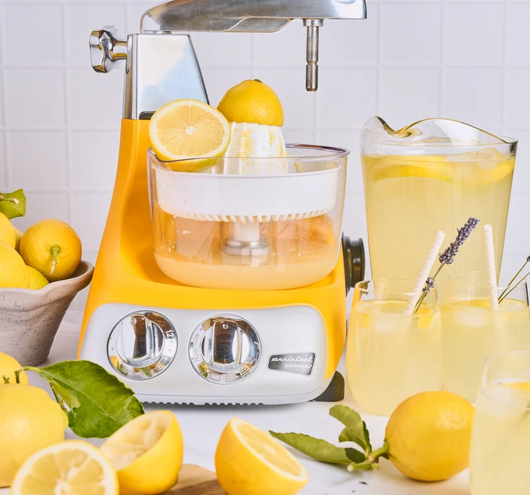 Ankarsrum Assistent Original sunbeam yellow stand mixer with citrus juicer attachment squeezing fresh lemons beside lemonade pitcher.