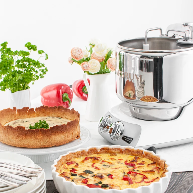 Ankarsrum Original stand mixer beside savory vegetable quiche and herb tart, styled with fresh flowers and red peppers.