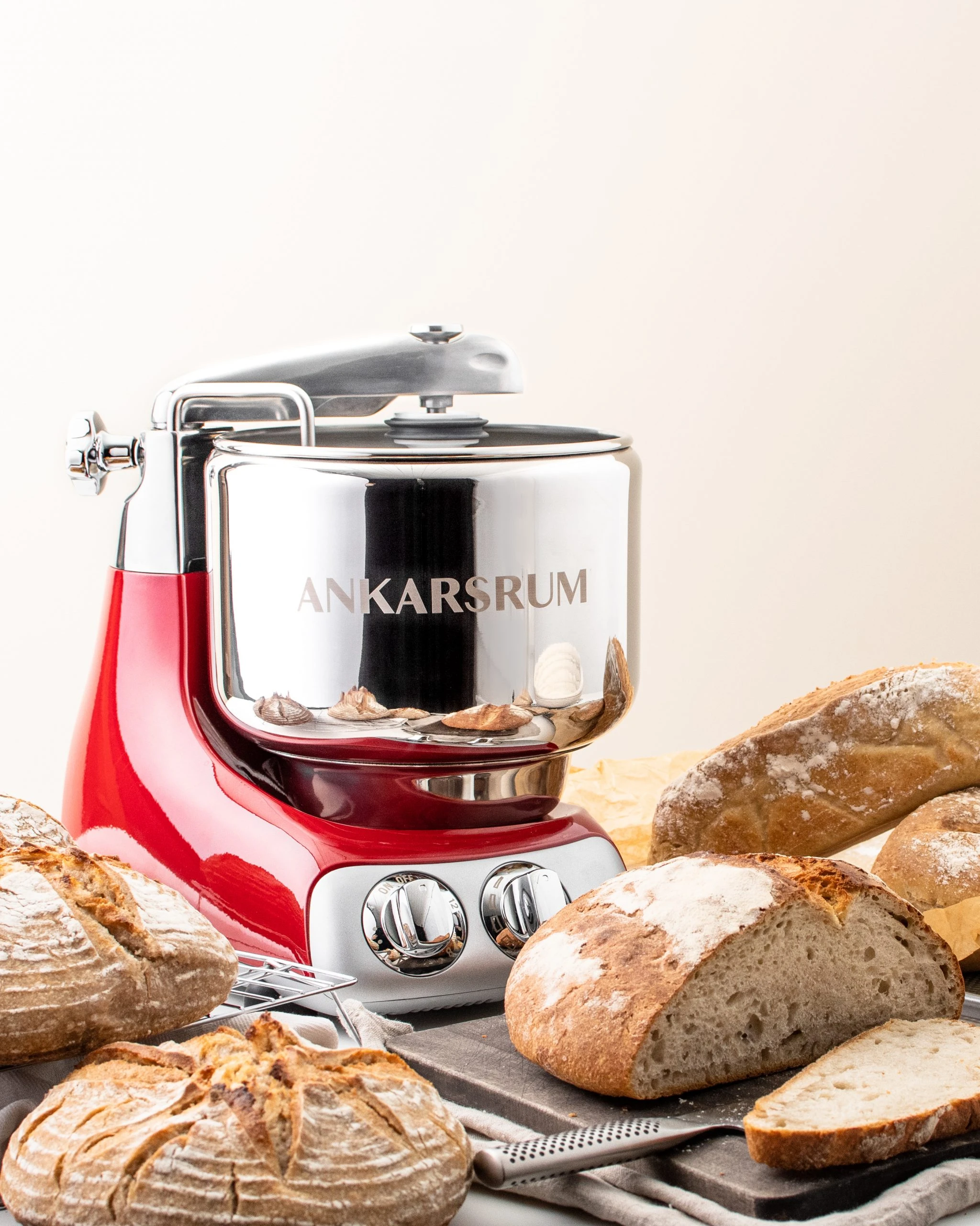 Ankarsrum red stand mixer with chrome bowl surrounded by freshly baked artisan sourdough bread loaves on a dark board.