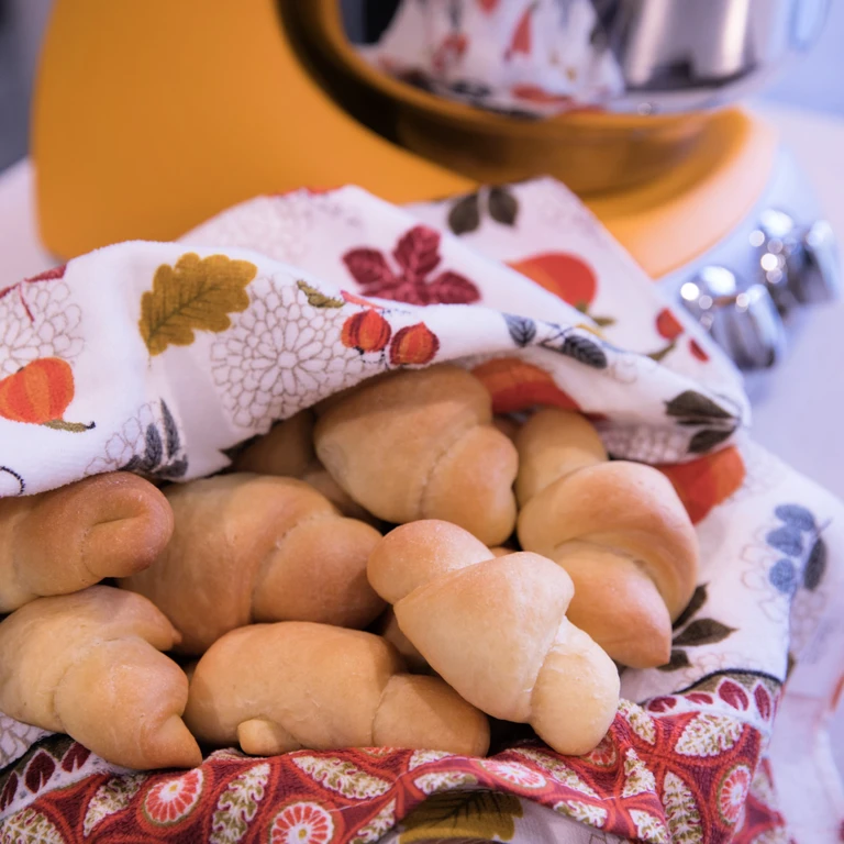 Freshly baked crescent rolls in a floral cloth basket beside an Ankarsrum stand mixer in a cozy kitchen setting.