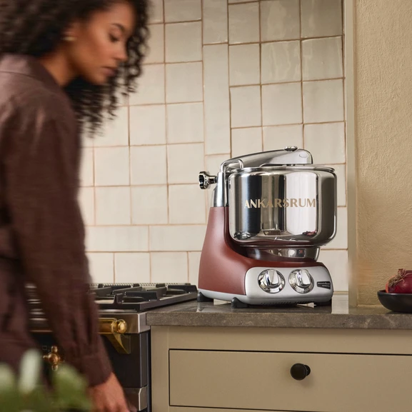 Ankarsrum Assistent Original in Rustic Maroon on a kitchen countertop in a bright kitchen, with a person partially visible in the image.