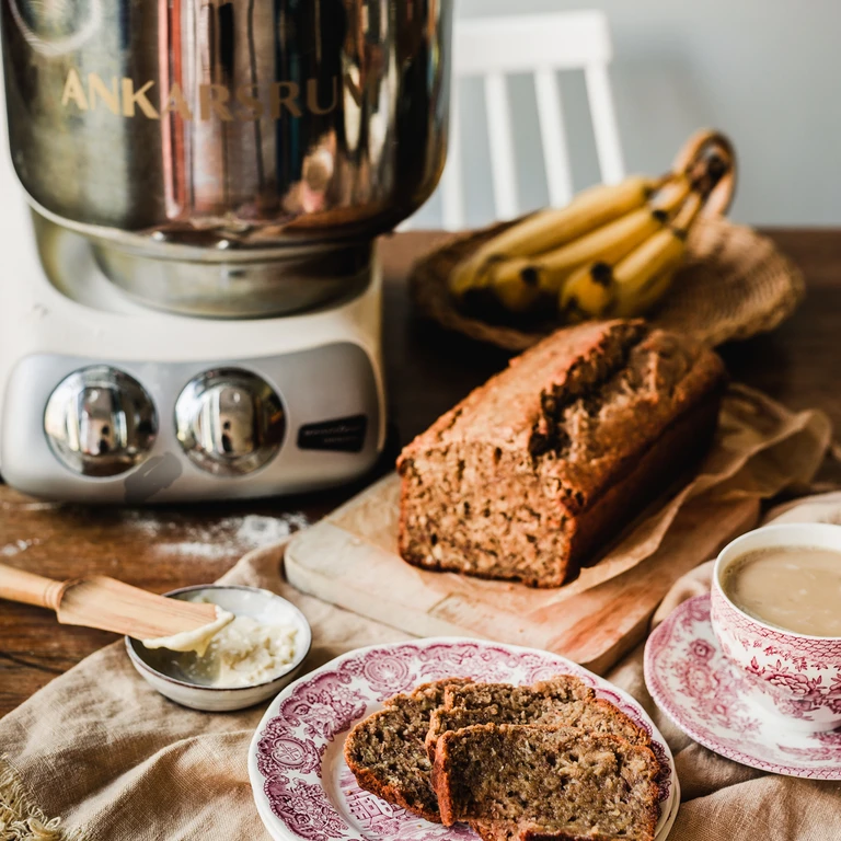 Ankarsrum stand mixer beside freshly baked banana bread loaf, sliced on decorative plate with butter and coffee cup.
