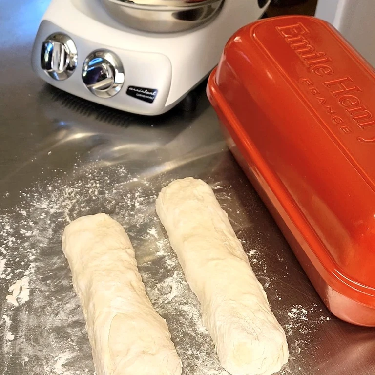 Two shaped bread dough logs on a floured surface beside an Emile Henry France loaf pan and Ankarsrum stand mixer.