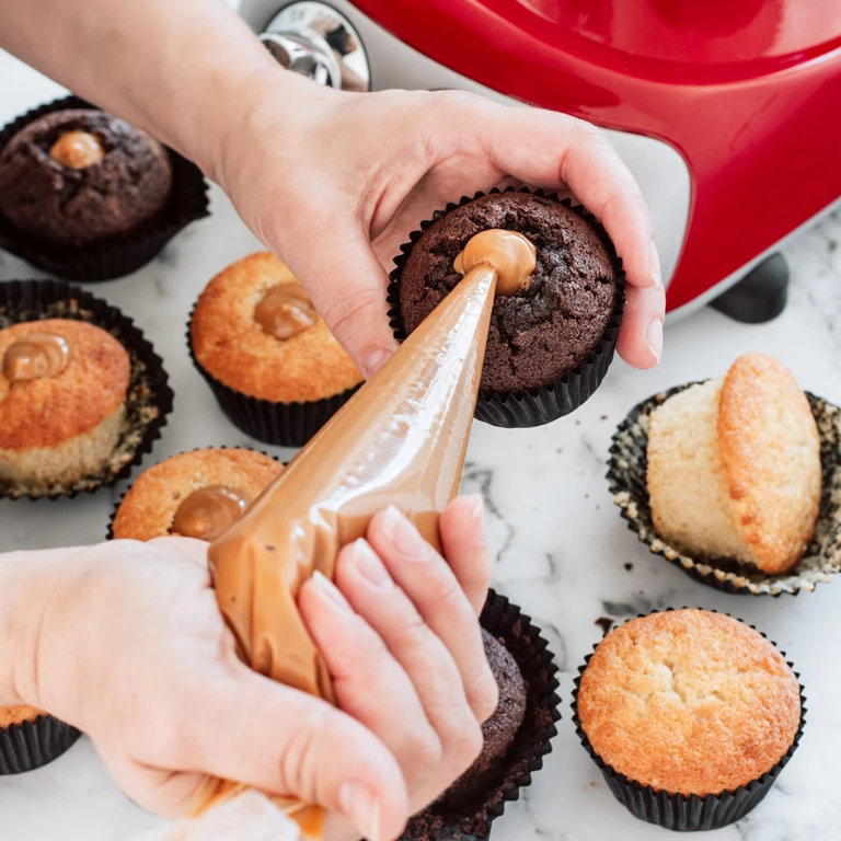 Hands filling chocolate and vanilla cupcakes with caramel using a piping bag beside a red Ankarsrum stand mixer.
