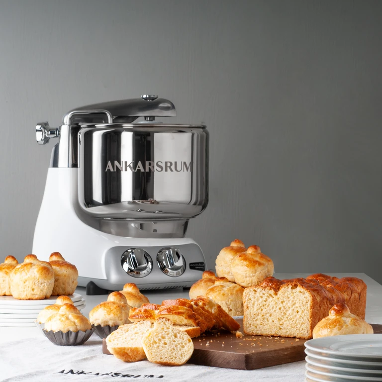 Ankarsrum stand mixer in white and chrome surrounded by freshly baked brioche rolls and sliced loaf bread on a table.