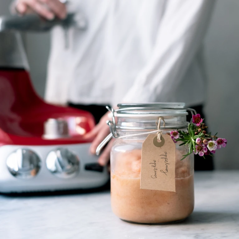 Jar of homemade applesauce with a kraft tag labeled Savodia applesauce, decorated with pink flowers, beside a red Ankarsrum mixer.