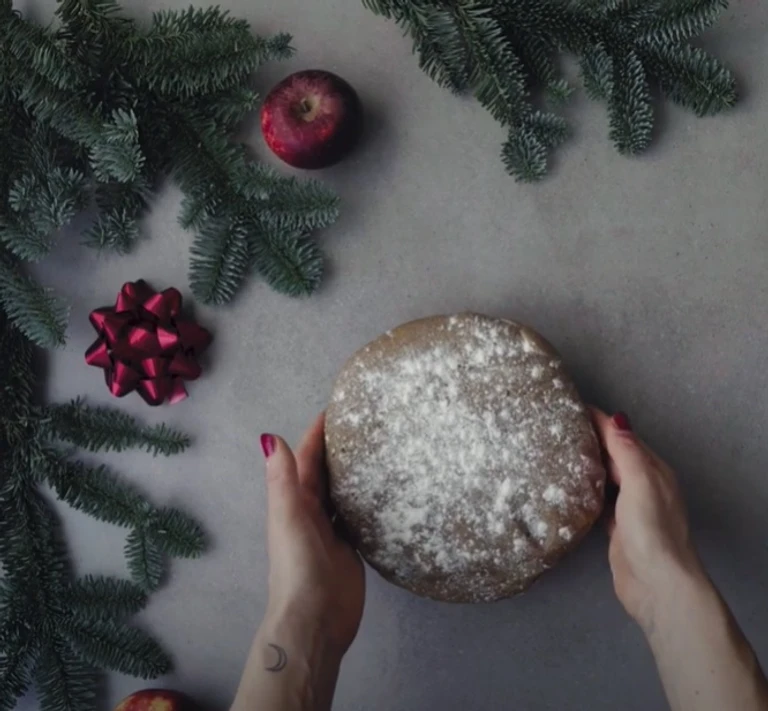 Hands holding a round flour-dusted dough ball, surrounded by pine branches, red apples, and a gift bow on a grey surface.