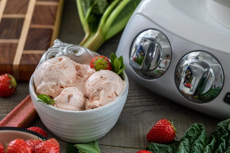 Strawberry rhubarb ice cream in a white bowl beside an Ankarsrum stand mixer, surrounded by fresh strawberries and rhubarb.