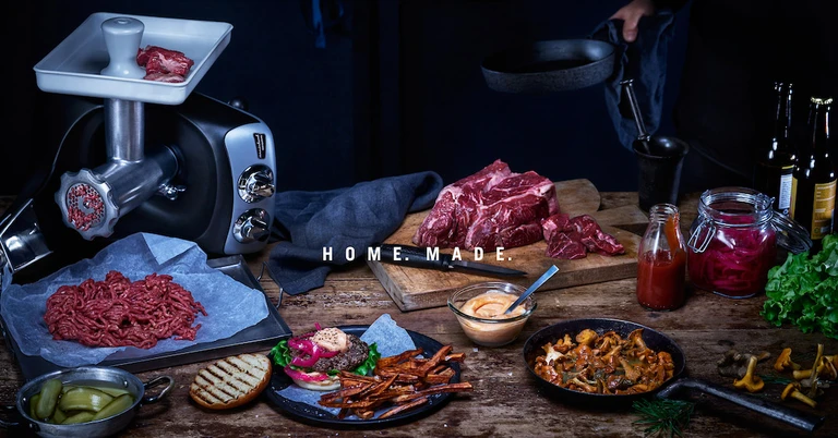 Home. Made. Ankarsrum meat grinder with fresh ground beef, raw cuts, burger, fries, sauces, and sautéed mushrooms on rustic table.