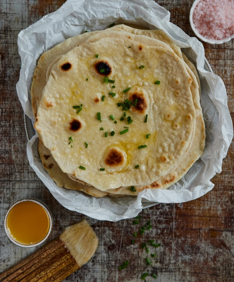 Stack of homemade flatbreads brushed with butter, topped with fresh chives, served in parchment paper with pink salt nearby.
