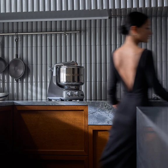 Ankarsrum Assistent Original in Black Chrome on a kitchen countertop, viewed from a distance, with a woman moving beside the machine in the kitchen.