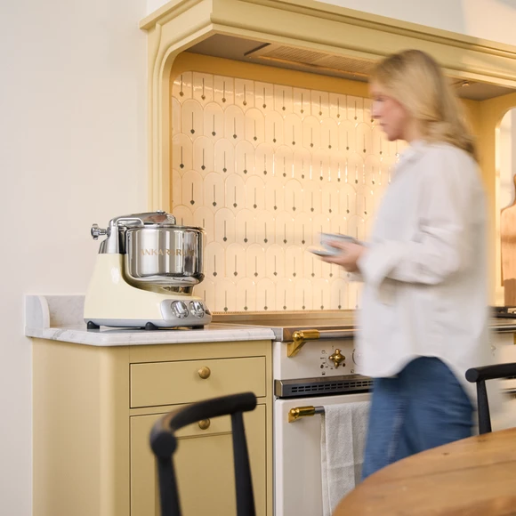 Ankarsrum Assistent Original in Creme Light on a light kitchen countertop in a yellow kitchen, with a person walking beside the countertop.