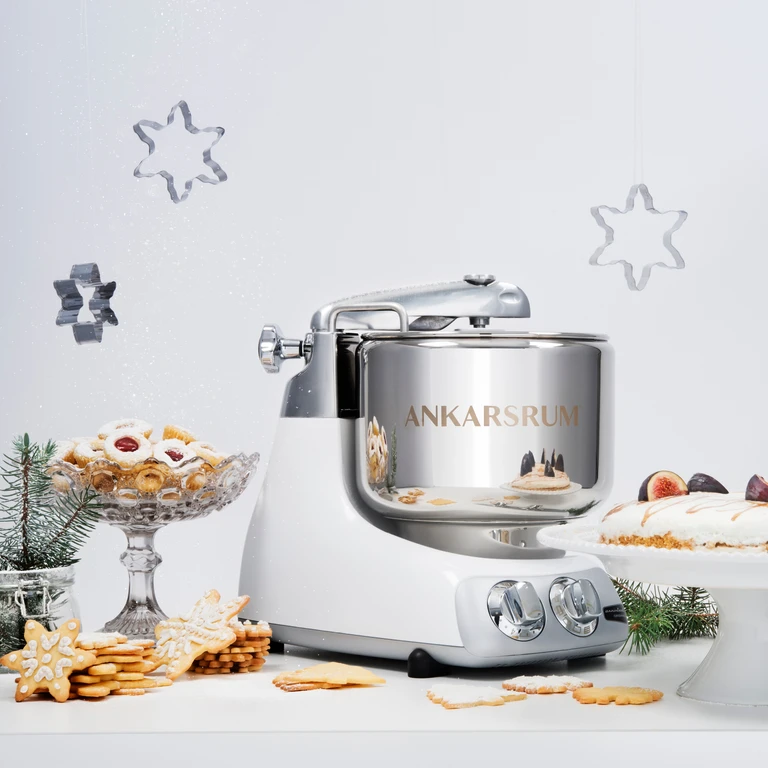 Ankarsrum stand mixer in white and chrome surrounded by Christmas cookies, gingerbread stars, and holiday baked goods.