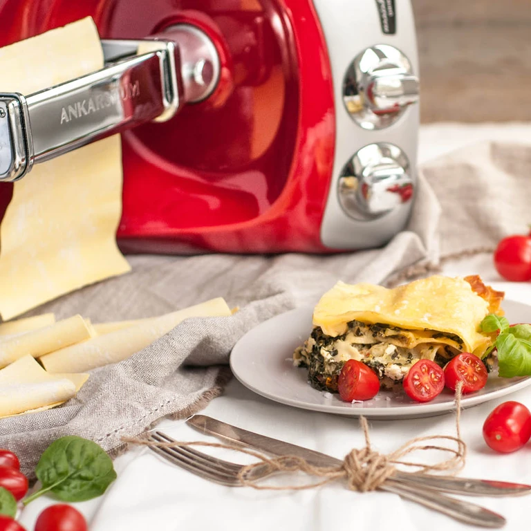 Ankarsrum red stand mixer with pasta roller attachment making fresh lasagna sheets, served with spinach filling and cherry tomatoes.