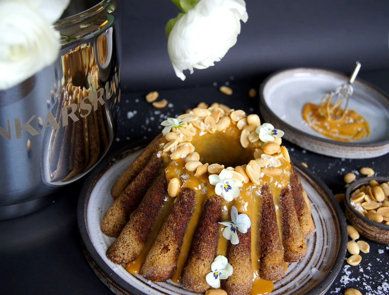 Ankarsrum bundt cake topped with caramel glaze, roasted peanuts, and edible flowers on a rustic ceramic plate.
