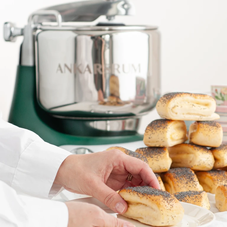 Hands arranging poppy seed rolls beside a green Ankarsrum stand mixer on a white surface.