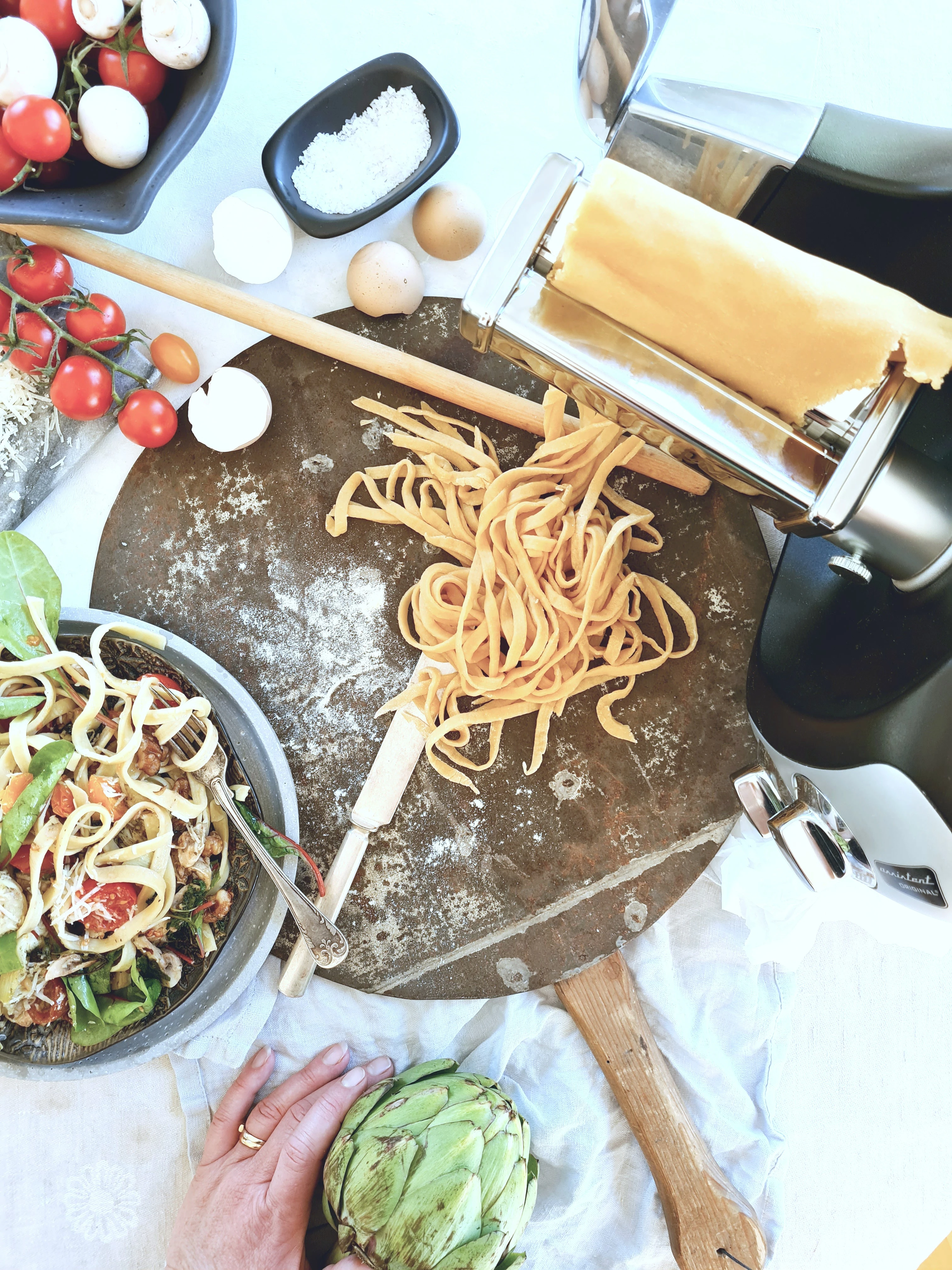 Freshly made tagliatelle pasta on a floured board beside an Ankarsrum stand mixer with pasta roller attachment, eggs, and tomatoes.