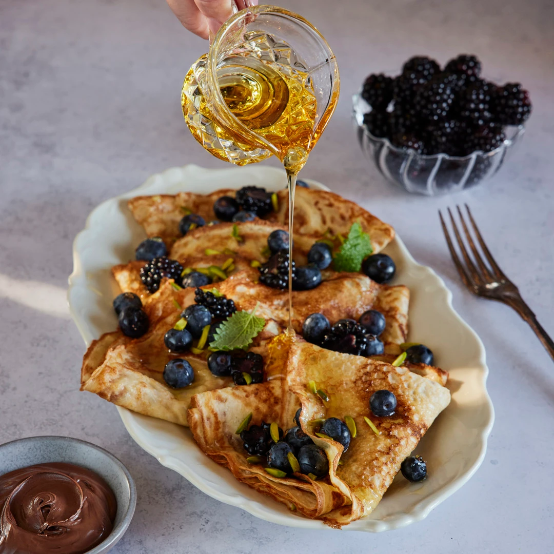 Honung hälls över vikta crêpes toppade med blåbär, björnbär och pistagenötter på ett serveringsfat. En skål med björnbär och en skål med chokladkräm står bredvid.