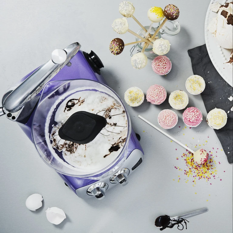 Ankarsrum purple stand mixer with whipped cream batter, surrounded by decorated cake pops and frosted cupcakes with sprinkles.