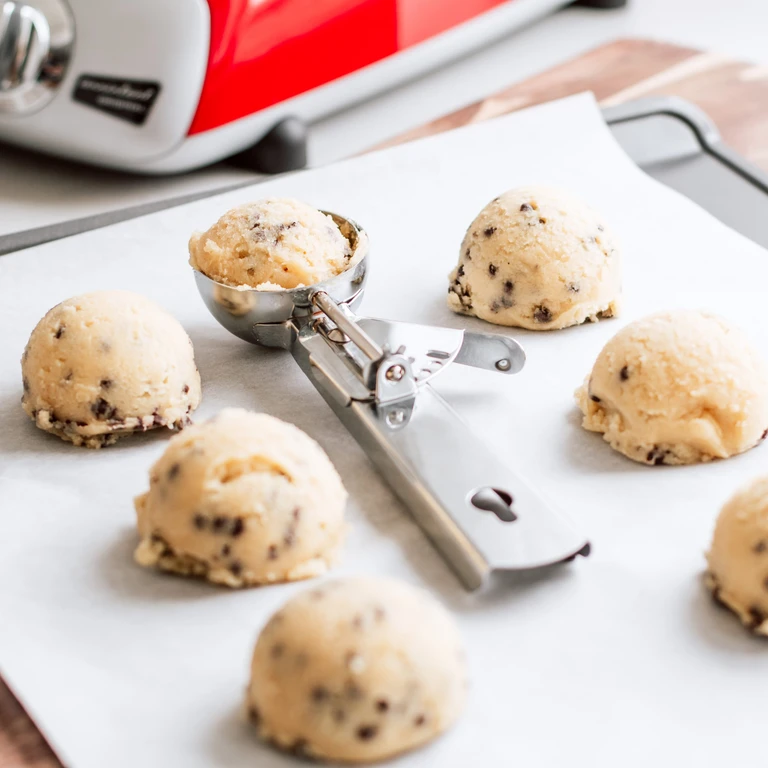 Chocolate chip cookie dough balls on parchment paper with a stainless steel scoop and red Ankarsrum stand mixer in background.