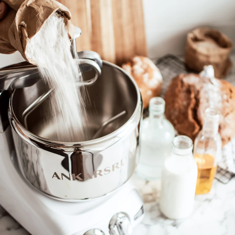 Flour being poured into an Ankarsrum stainless steel stand mixer bowl, with bread loaves and milk bottles in the background.