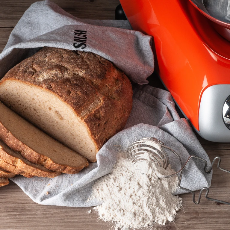 Sliced homemade bread loaf on Ankarsrum linen cloth with spilled flour and red Ankarsrum stand mixer in background.