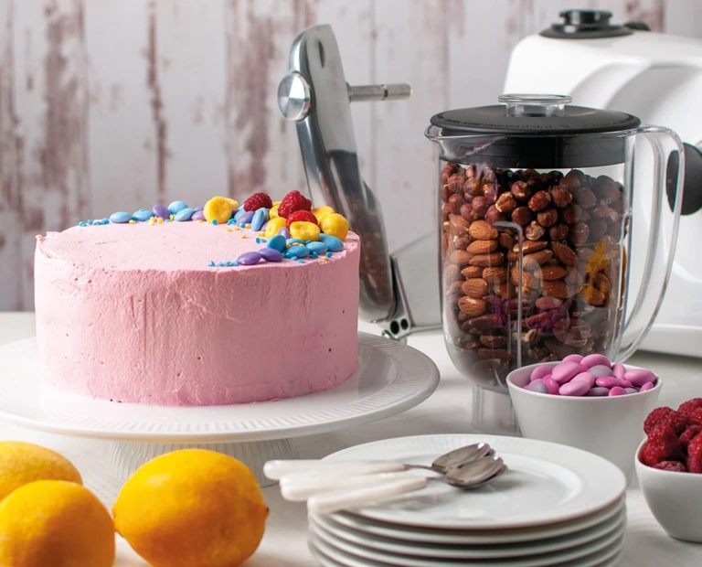 Pink frosted cake with colorful candy toppings beside an Ankarsrum blender filled with mixed nuts.