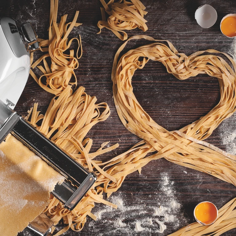 Fresh homemade fettuccine pasta arranged in a heart shape beside an Ankarsrum pasta roller machine with egg yolks and flour.