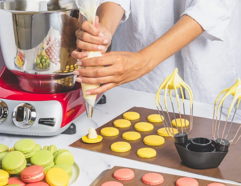 Hands piping yellow cream filling onto macaron shells using an Ankarsrum Original red stand mixer with whisk attachment nearby.