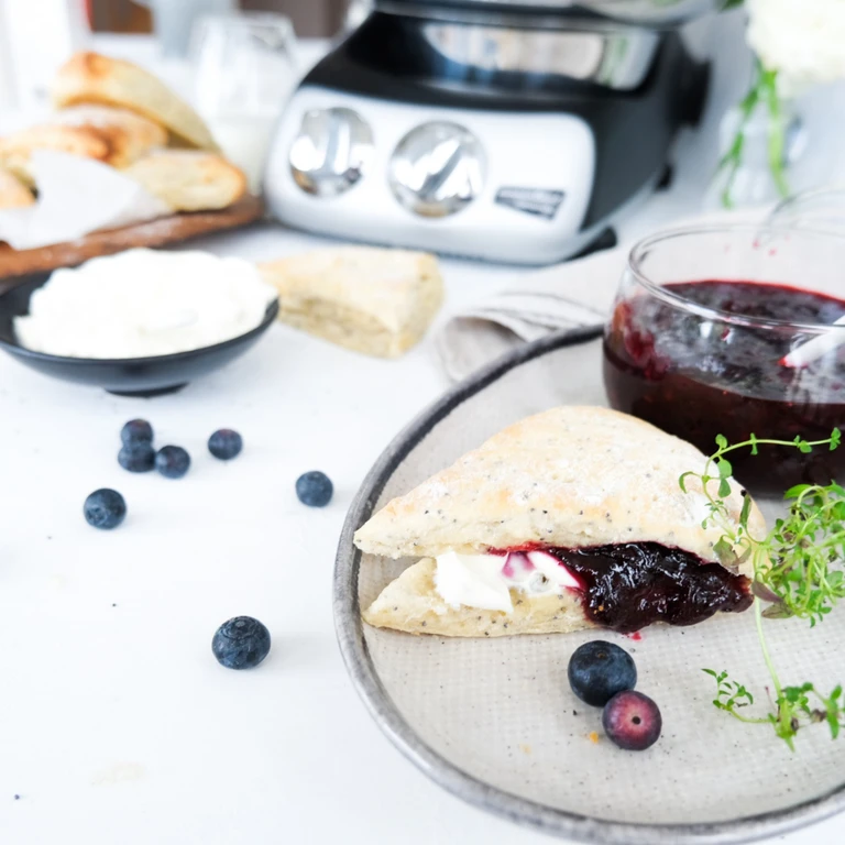 Scone with cream and blueberry jam on a plate, fresh blueberries, and Ankarsrum stand mixer in background.