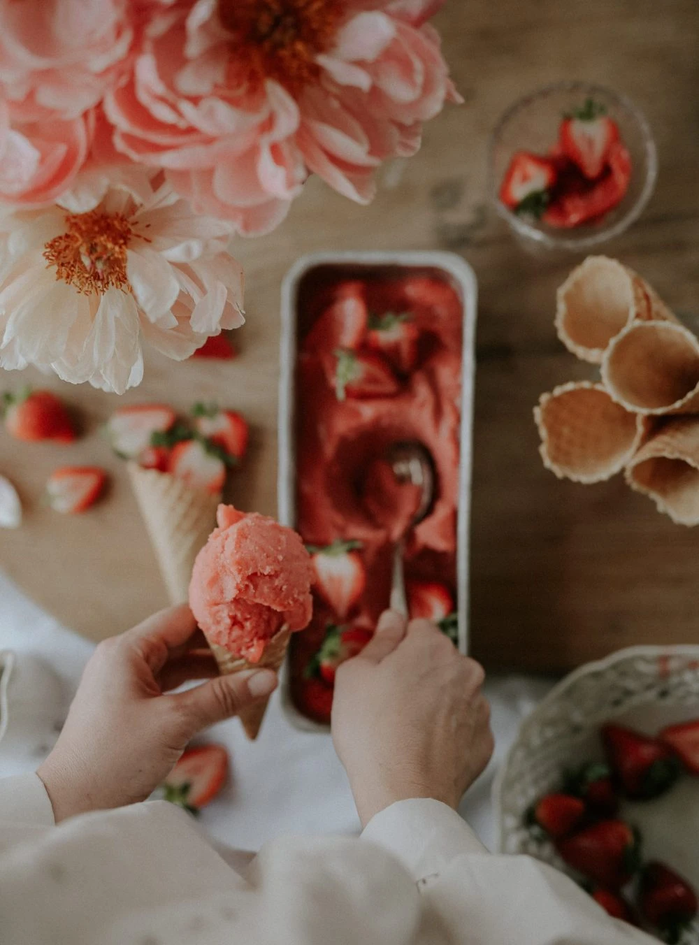 Hands holding a waffle cone with strawberry sorbet scoop, fresh strawberries, and pink peonies on wooden table.