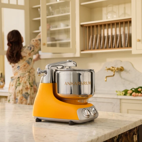 Ankarsrum Assistent Original in Sunbeam Yellow on a marble kitchen island in a bright kitchen, with a person putting items away in a kitchen cabinet in the background.