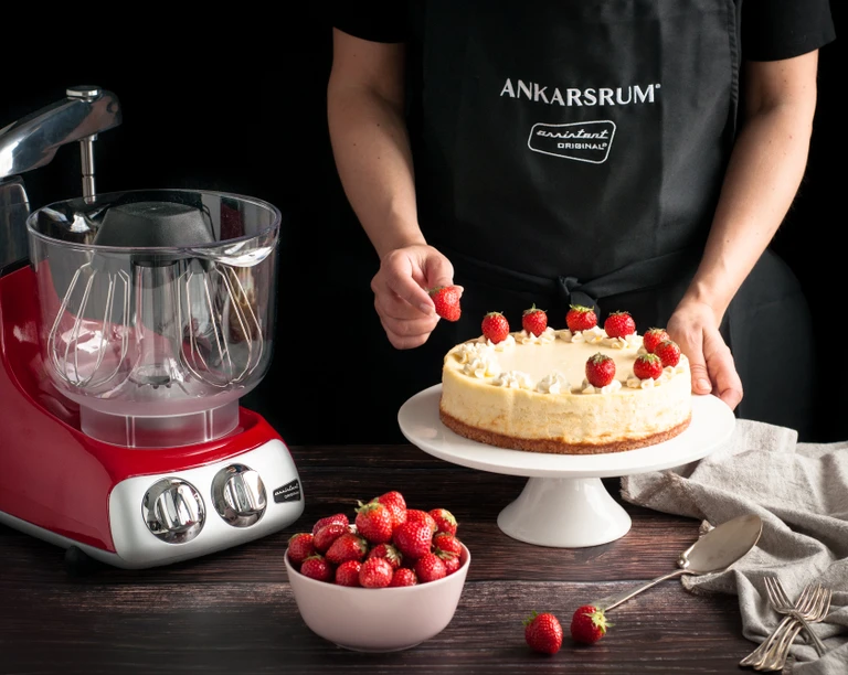 Person decorating a cheesecake with strawberries beside a red Ankarsrum Assistent Original stand mixer.