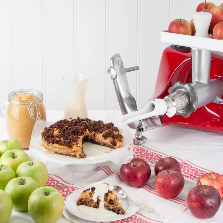 Ankarsrum red stand mixer with grinder attachment, apple pie, fresh red and green apples, cream, and applesauce on white table.