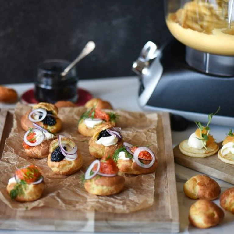 Savory choux pastry bites topped with smoked salmon, cream cheese, caviar, and red onion on parchment, with Ankarsrum mixer in background.