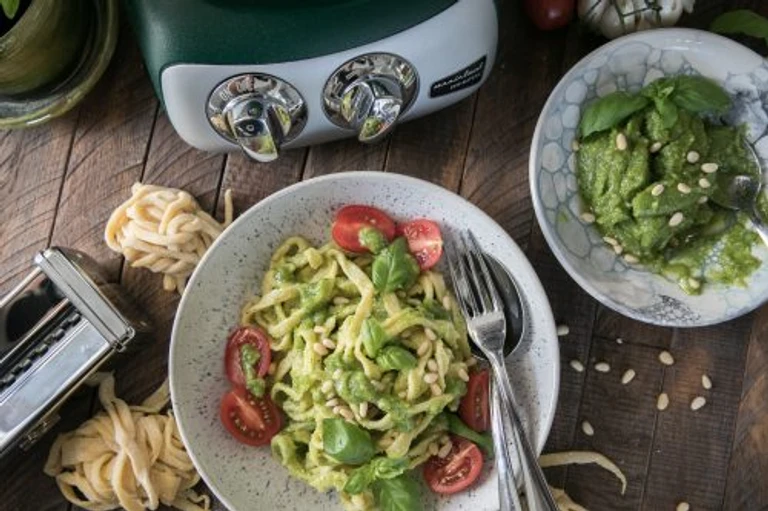 Ankarsrum stand mixer beside homemade pasta with pesto, cherry tomatoes, basil, and pine nuts on a rustic wooden table.