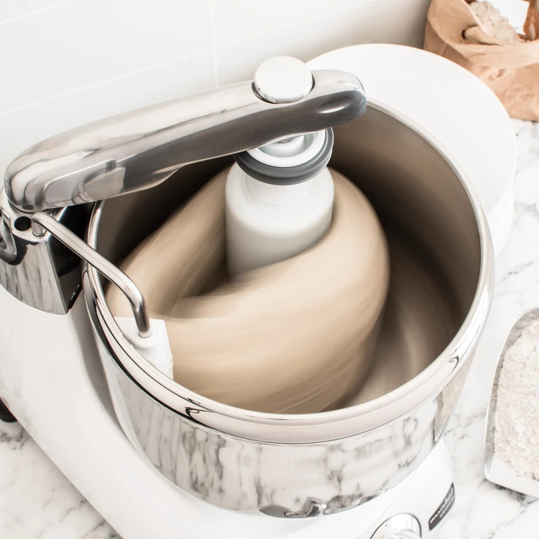 Ankarsrum stand mixer kneading bread dough in polished stainless steel bowl with dough roller attachment in motion.