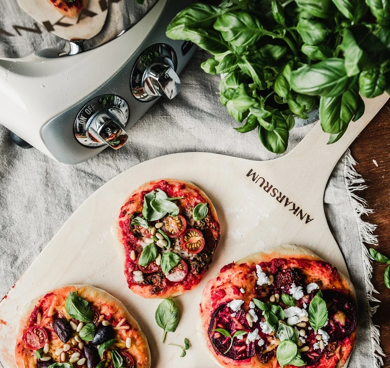 Ankarsrum pizza peel board with three mini pizzas topped with basil, tomatoes, beets, and pine nuts beside a stand mixer.