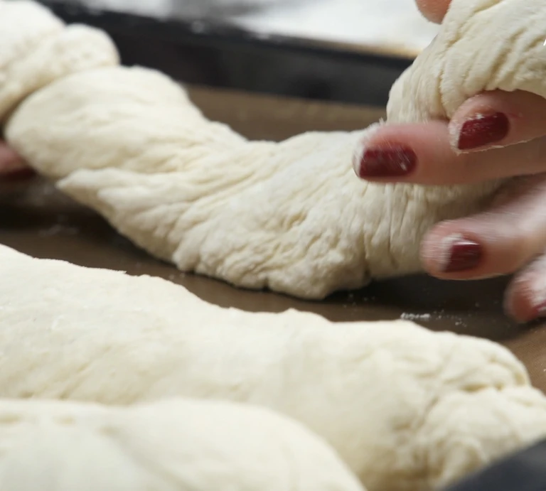 Hands with red nails shaping white bread dough strands on a baking tray using an Ankarsrum stand mixer.